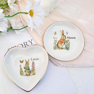 Puede incluir: Dos platos decorativos para baratijas. Uno tiene forma de corazón, el otro es redondo. Ambos son blancos con bordes dorados y presentan una ilustración de acuarela de un conejo con una chaqueta. El plato en forma de corazón tiene el nombre "Lucas" y el plato redondo tiene el nombre "Mason".