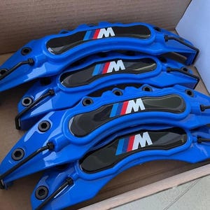 Puede incluir: Cuatro pinzas de freno azules con logotipos "M" negros y rojos. Las pinzas están diseñadas para adaptarse a un vehículo BMW.