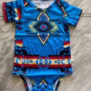 May include: A blue baby bodysuit with a geometric pattern in red, yellow, black, and white. The short-sleeved onesie features a round neckline and snap closures at the bottom. The design has a Native American inspired motif.