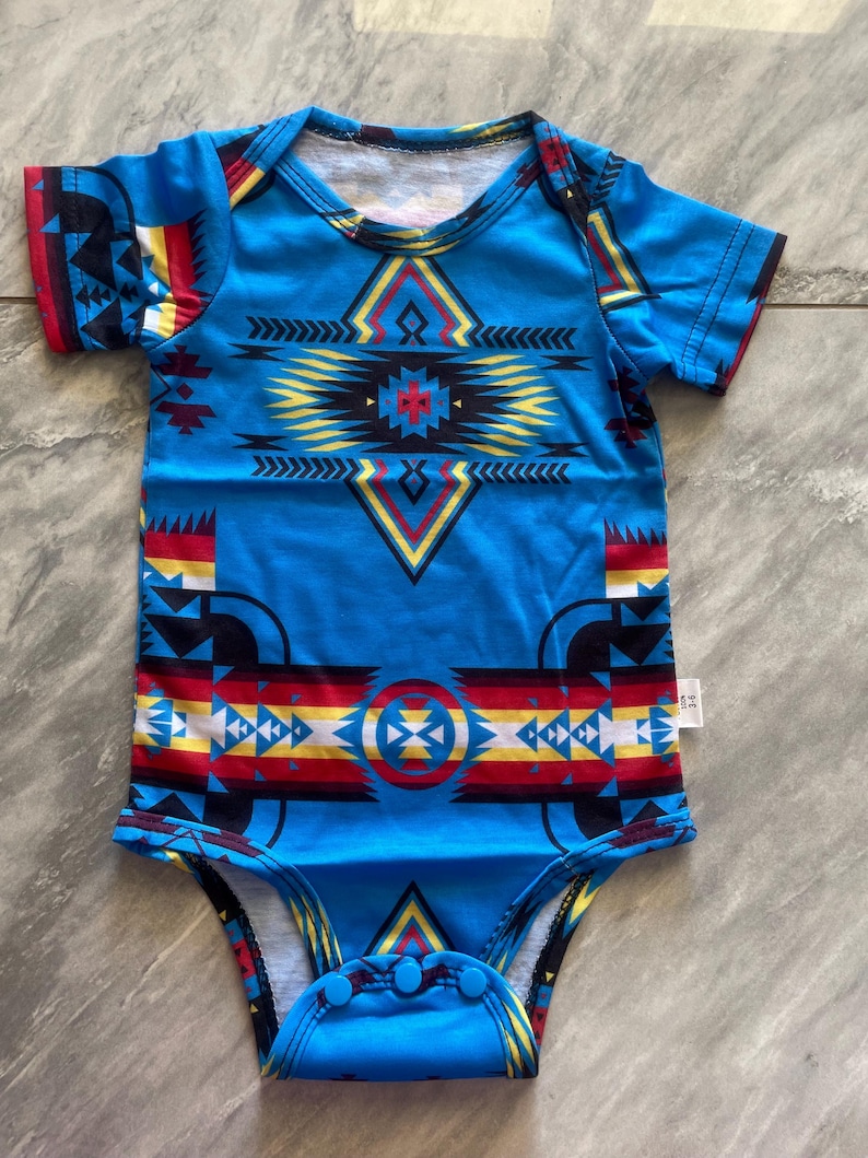 May include: A blue baby bodysuit with a geometric pattern in red, yellow, black, and white. The short-sleeved onesie features a round neckline and snap closures at the bottom. The design has a Native American inspired motif.