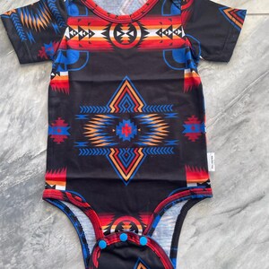 May include: Black baby bodysuit with a colorful geometric pattern in red, blue, orange, and white. The bodysuit has short sleeves and snap closures at the bottom. The word "Black" is printed at the top.