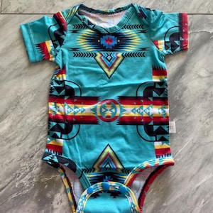 May include: A turquoise baby bodysuit with a colorful geometric pattern. The design includes red, yellow, black, and white shapes. The short-sleeved onesie has snap closures at the bottom.