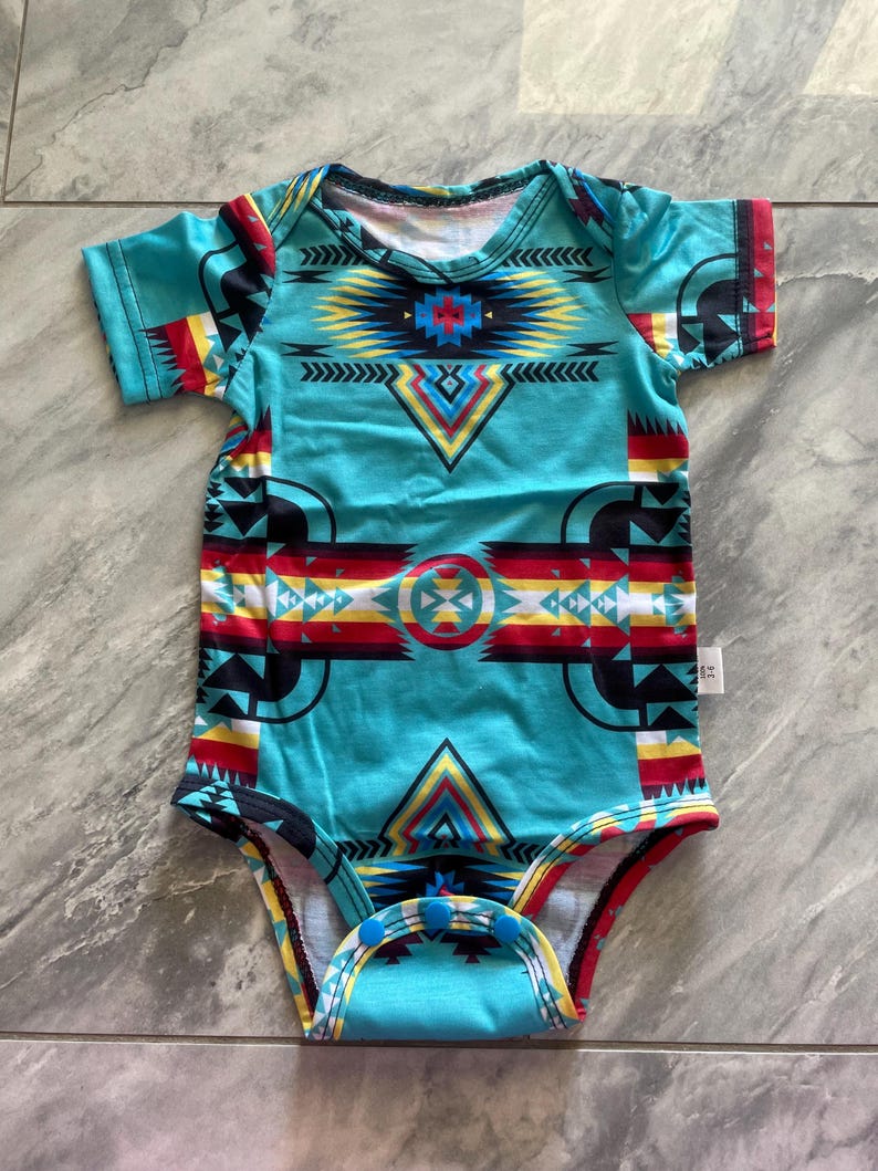 May include: A turquoise baby bodysuit with a colorful geometric pattern. The design includes red, yellow, black, and white shapes. The short-sleeved onesie has snap closures at the bottom.