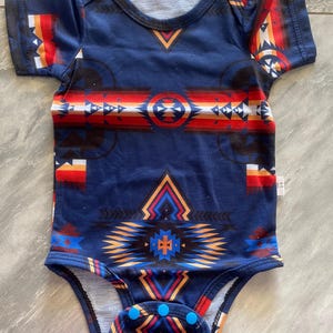 May include: A navy blue baby onesie with a Southwestern-inspired geometric pattern in red, orange, white, and black. The short-sleeved bodysuit has a round neckline and snap closures at the bottom.