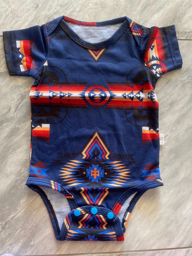 May include: A navy blue baby onesie with a Southwestern-inspired geometric pattern in red, orange, white, and black. The short-sleeved bodysuit has a round neckline and snap closures at the bottom.
