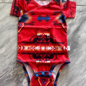 May include: Red baby bodysuit with a Southwestern-inspired geometric pattern in black, blue, yellow, and white. The short-sleeved onesie has blue snap closures at the bottom and a white tag.