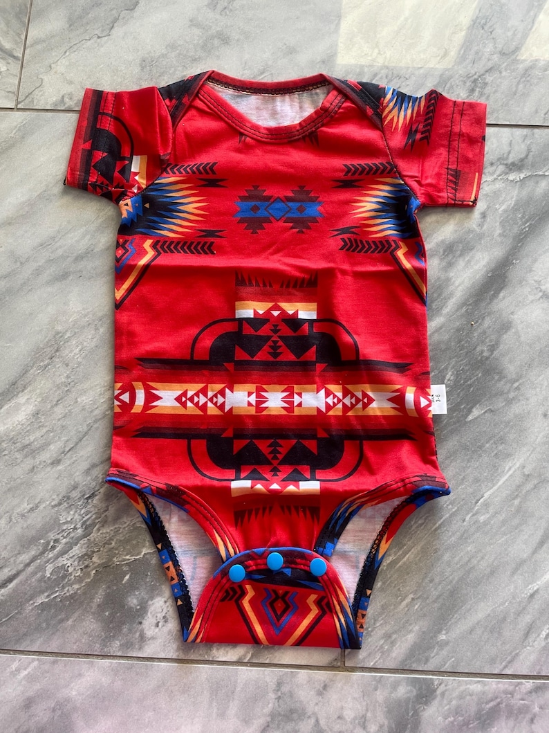 May include: Red baby bodysuit with a Southwestern-inspired geometric pattern in black, blue, yellow, and white. The short-sleeved onesie has blue snap closures at the bottom and a white tag.