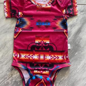 May include: A red baby onesie with a Southwestern-inspired geometric pattern in shades of black, blue, orange, and white. The short-sleeved bodysuit has a round neckline and blue snap closures at the bottom.