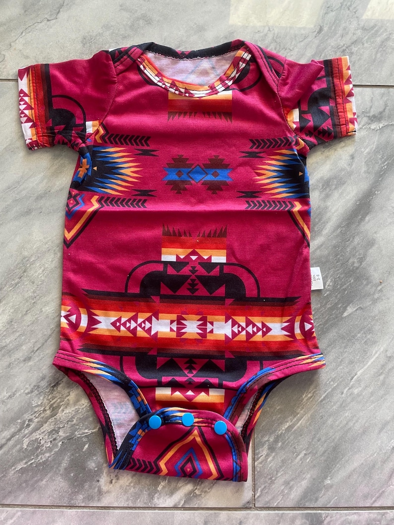 May include: A red baby onesie with a Southwestern-inspired geometric pattern in shades of black, blue, orange, and white. The short-sleeved bodysuit has a round neckline and blue snap closures at the bottom.