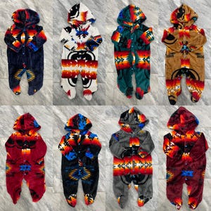 May include: Eight baby onesies with different geometric patterns in various colors, including red, blue, green, brown, and white. Each onesie has a hood with a pointed tip and a button closure.