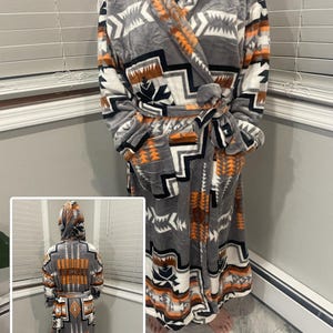May include: A hooded robe with a Southwestern pattern in grey, white, orange, and black. The robe has a tie belt and is made of a soft, plush material. The robe is displayed indoors.