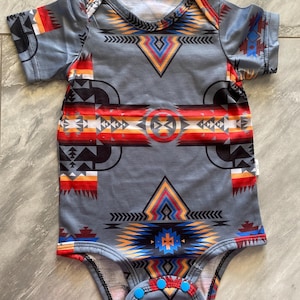 May include: Gray baby bodysuit with a Southwestern-inspired geometric pattern in shades of red, orange, yellow, and blue. The short-sleeved onesie has blue snap closures at the bottom and a white neckline.