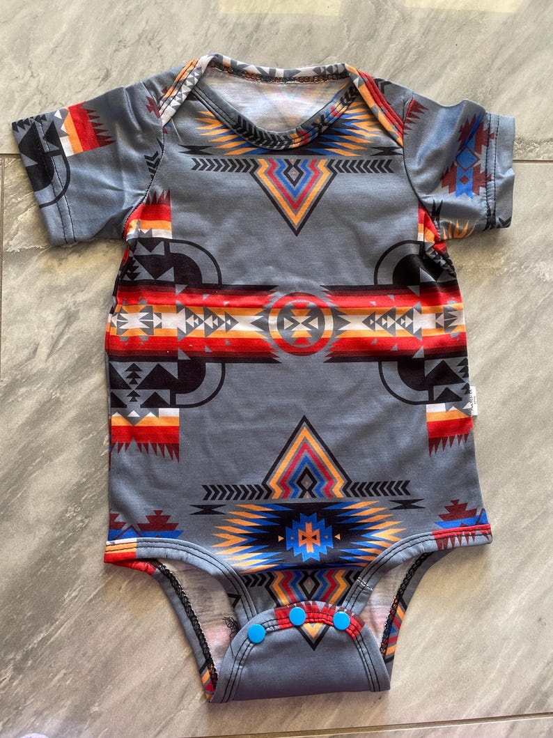 May include: Gray baby bodysuit with a Southwestern-inspired geometric pattern in shades of red, orange, yellow, and blue. The short-sleeved onesie has blue snap closures at the bottom and a white neckline.