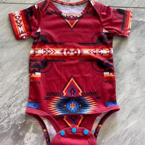 May include: Red baby bodysuit with a Southwestern-inspired geometric pattern in black, white, orange, and blue. The short-sleeved onesie has a V-neck and three blue snaps at the bottom.