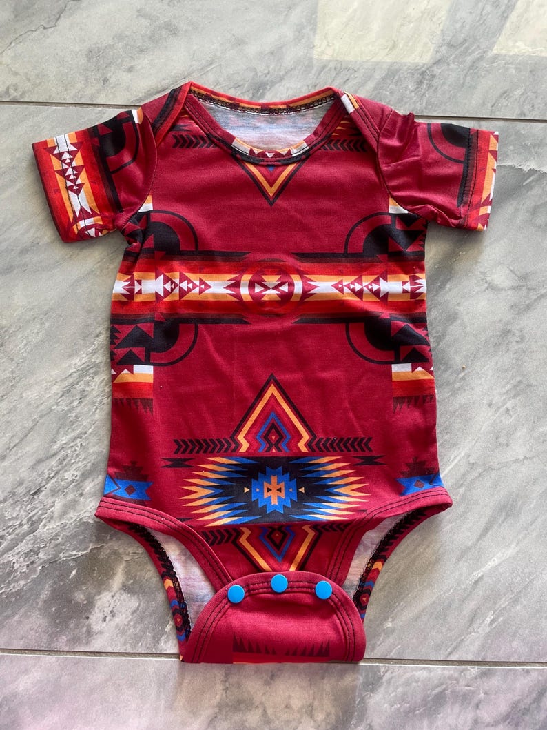 May include: Red baby bodysuit with a Southwestern-inspired geometric pattern in black, white, orange, and blue. The short-sleeved onesie has a V-neck and three blue snaps at the bottom.