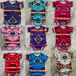 May include: Nine baby bodysuits with short sleeves and a colorful geometric pattern. The bodysuits are in various colors, including navy blue, turquoise, pink, purple, red, and white. Each bodysuit has a unique design with a Southwestern or Native American-inspired motif.