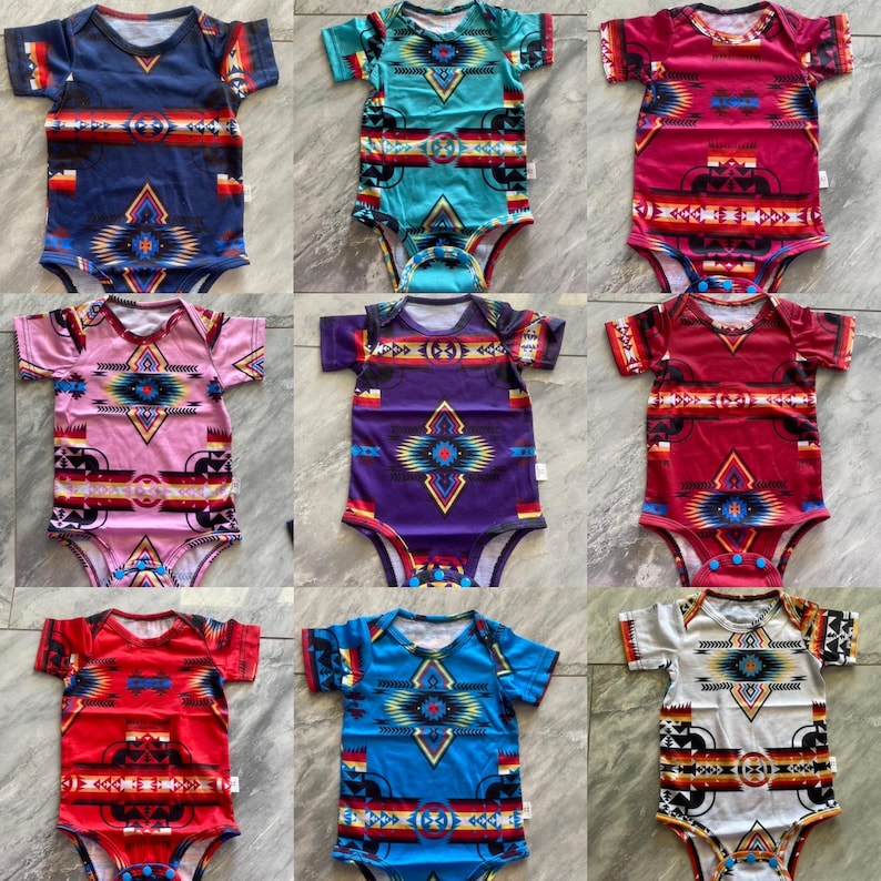 May include: Nine baby bodysuits with short sleeves and a colorful geometric pattern. The bodysuits are in various colors, including navy blue, turquoise, pink, purple, red, and white. Each bodysuit has a unique design with a Southwestern or Native American-inspired motif.