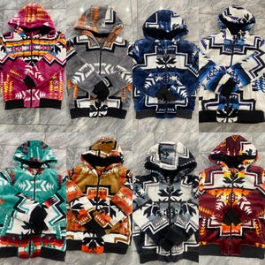 May include: A collection of hooded zip-up jackets with a Southwestern-inspired geometric pattern. The jackets feature a variety of colors, including pink, gray, blue, teal, brown, and burgundy. Each jacket has a black ribbed hem and cuffs.