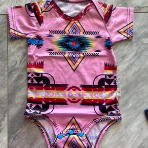 May include: Pink baby bodysuit with a colorful geometric pattern. The design features a central diamond shape with blue, yellow, and red accents, and a tribal-inspired motif. The bodysuit has short sleeves and blue snap closures.