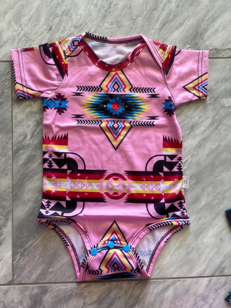 May include: Pink baby bodysuit with a colorful geometric pattern. The design features a central diamond shape with blue, yellow, and red accents, and a tribal-inspired motif. The bodysuit has short sleeves and blue snap closures.