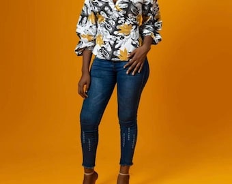 May include: A woman wearing a white and black wrap top with a yellow floral print and blue jeans. She is standing on a yellow background.