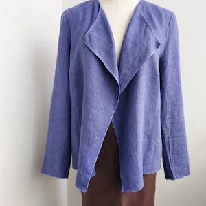 May include: A periwinkle blue linen jacket with a shawl collar and long sleeves. The jacket is displayed on a mannequin, paired with a brown leather skirt. The jacket has a relaxed, open front design.