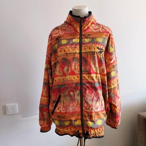 Op de afbeelding: Een vintage Reebok-jack met een kleurrijke paisley-print in tinten rood, oranje, geel en blauw. Het jack heeft een ritssluiting en een trekkoord aan de onderkant.