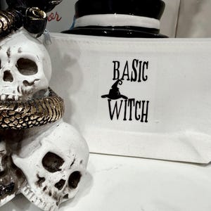Könnte beinhalten: Weiße Kosmetiktasche aus Canvas mit dem Text "BASIC WITCH" in Schwarz, mit einer Hexenhut-Grafik. Eine dekorative Totenkopf-Statue mit einer goldenen Schlange im Vordergrund.