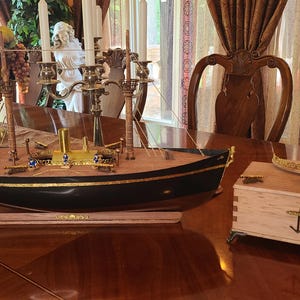 Könnte beinhalten: Ein hölzernes Schiffsmodell mit goldenen Akzenten und einem schwarzen Rumpf, auf einem Holzsockel ausgestellt. Das Schiff steht auf einem Tisch mit einer Holzkiste mit einem goldenen Anker auf dem Deckel.