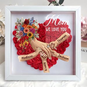 May include: A framed artwork featuring a heart made of red roses, with a bouquet of colorful flowers on the left. The words "Mom WE LOVE YOU" are displayed above a depiction of hands holding each other, with names Madison, Nancy, Thomas, and Jackson.