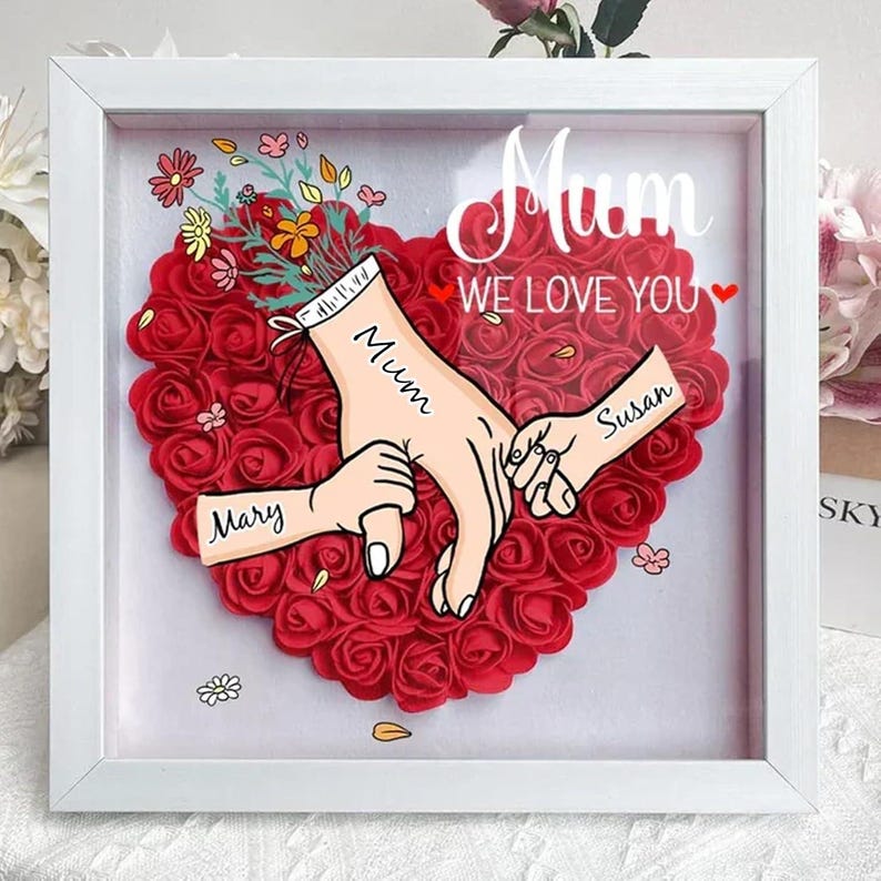 May include: White frame with a red heart made of roses. The heart has three hands holding each other with the names Mary, Mum, and Susan written on them. The text "Mum We Love You" is written above the heart.