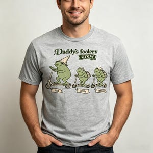 Puede incluir: Camiseta gris claro con una ilustración de dibujos animados de tres ranas verdes en patinetes. El texto "Daddy's foolery crew" está encima de las ranas, con los nombres "Dad", "Emily" y "James" debajo.