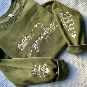 May include: Olive green sweatshirt with white embroidered text reading "mom" and "grandma" with dates and names on the sleeves. The sweatshirt has a crew neck and ribbed cuffs. The names include Olivia, Hannah, Rachel, Thomas, Max, Ella, Chris, Kevin, and Lindsey.