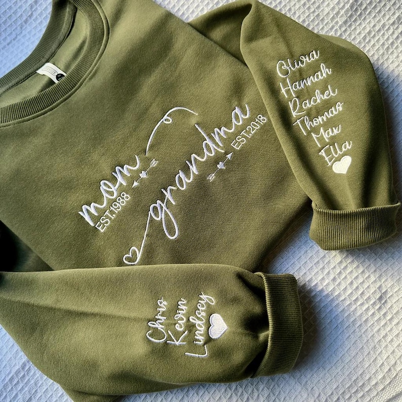 May include: Olive green sweatshirt with white embroidered text reading "mom" and "grandma" with dates and names on the sleeves. The sweatshirt has a crew neck and ribbed cuffs. The names include Olivia, Hannah, Rachel, Thomas, Max, Ella, Chris, Kevin, and Lindsey.