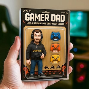 May include: A Gamer Dad figurine in a clear plastic case. The figurine depicts a man wearing a black hoodie and headphones, holding a game controller. The case includes three colorful game controllers with names: Henry, Sophia, and Emma. The text on the case reads "Gamer Dad."