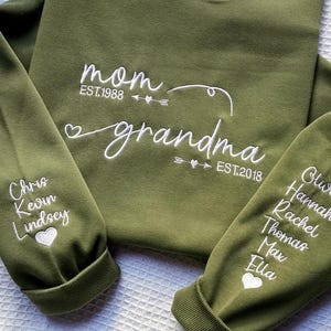 May include: An olive green sweatshirt with white embroidered text. The text on the front reads "mom EST.1988" and "grandma EST.2018". The sleeves display names and heart symbols.