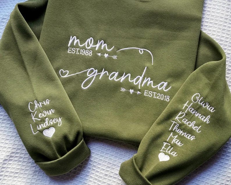 May include: An olive green sweatshirt with white embroidered text. The text on the front reads "mom EST.1988" and "grandma EST.2018". The sleeves display names and heart symbols.