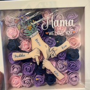 May include: A white frame with a floral arrangement of purple, pink, and blue roses. The frame has a clear plastic cover and the text "Mama We Love You" with the names "Freddie", "Estelle", "Evie", and "Mama" written on the roses.