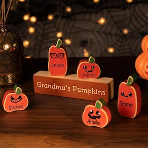 Pode incluir: Uma coleção de decorações de abóbora de madeira laranja com caules verdes. Cada abóbora tem um rosto e um nome diferentes, como "Lauren", "Aurora", "Michael", "Amelia" e "Emma". Uma placa de madeira diz "Grandma's Pumpkins".