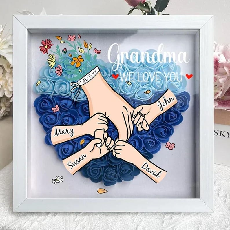 May include: A white frame with a blue and light blue heart made of artificial roses. The heart has four hands reaching into it, with the names Mary, Susan, John, and David written on the hands. The text "Grandma We Love You" is written above the heart.