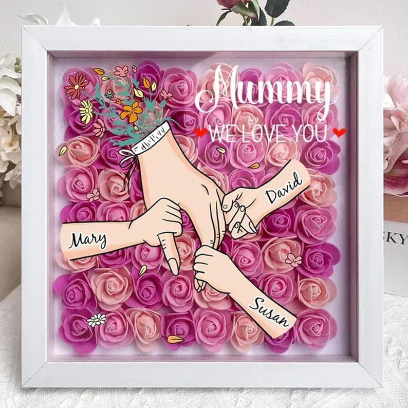 May include: A white frame with a pink and white floral background. The frame contains a drawing of three hands holding each other. The hands are labeled "Mary", "David", and "Susan". The text "Mummy We Love You" is written in pink letters at the top of the frame.