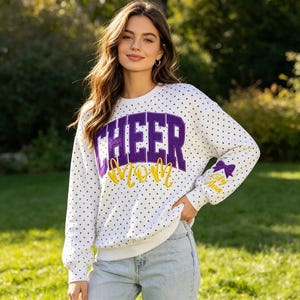 Peut inclure: Un sweat-shirt blanc à manches longues avec un motif à pois noirs. L'avant affiche le mot CHEER en grandes lettres violettes avec le mot MOM en écriture jaune en dessous. Un nœud violet et le nom Lily Ava figurent sur la manche.