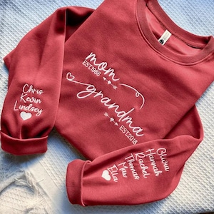 May include: A red sweatshirt with white embroidered text that reads "mom" and "grandma". The sleeves have names embroidered in white, with a heart symbol. The sweatshirt has a crew neck.