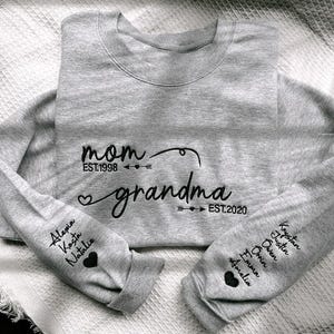 May include: A heather gray sweatshirt with the words "mom" and "grandma" in black script. The sleeves display names and hearts. The sweatshirt is a crew neck style.