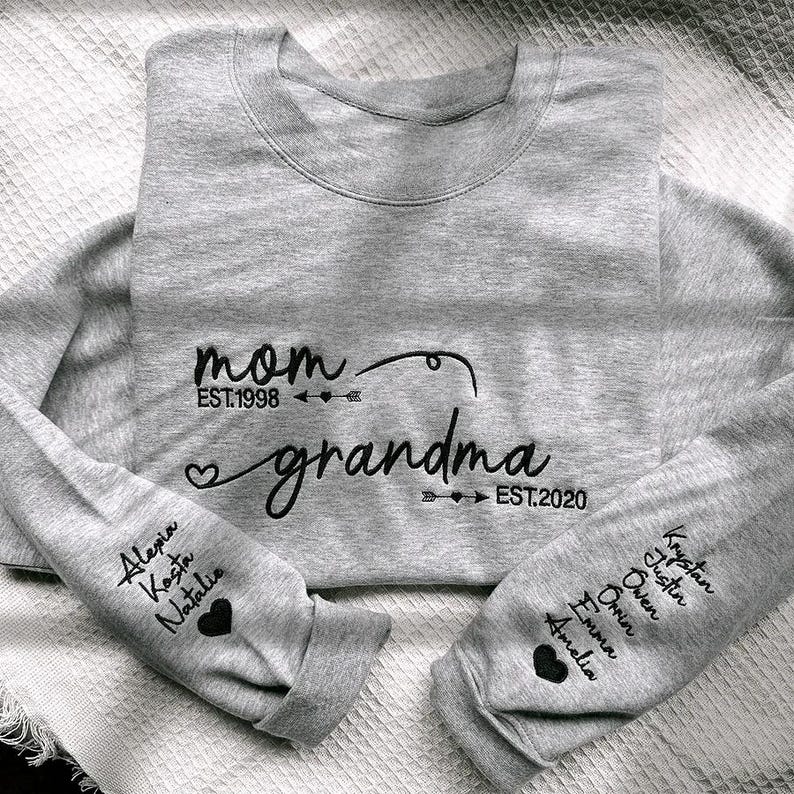 May include: A heather gray sweatshirt with the words "mom" and "grandma" in black script. The sleeves display names and hearts. The sweatshirt is a crew neck style.