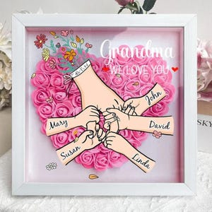 May include: A white frame with a pink heart-shaped background of artificial roses. The heart has five hands reaching out from it, with the names Mary, Susan, Linda, John, and David written on the arms. The text "Grandma We Love You" is written at the top of the frame.
