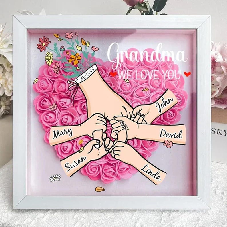 May include: A white frame with a pink heart-shaped background of artificial roses. The heart has five hands reaching out from it, with the names Mary, Susan, Linda, John, and David written on the arms. The text "Grandma We Love You" is written at the top of the frame.