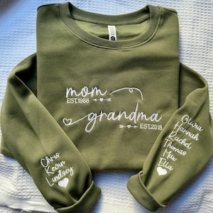May include: Olive green sweatshirt with white embroidered text reading "mom" and "grandma". The sleeves feature names and dates. A heart and arrow design is included. The sweatshirt is made of a soft material.