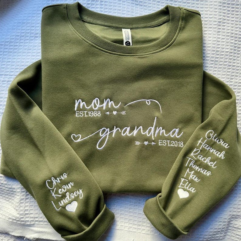 May include: Olive green sweatshirt with white embroidered text reading "mom" and "grandma". The sleeves feature names and dates. A heart and arrow design is included. The sweatshirt is made of a soft material.