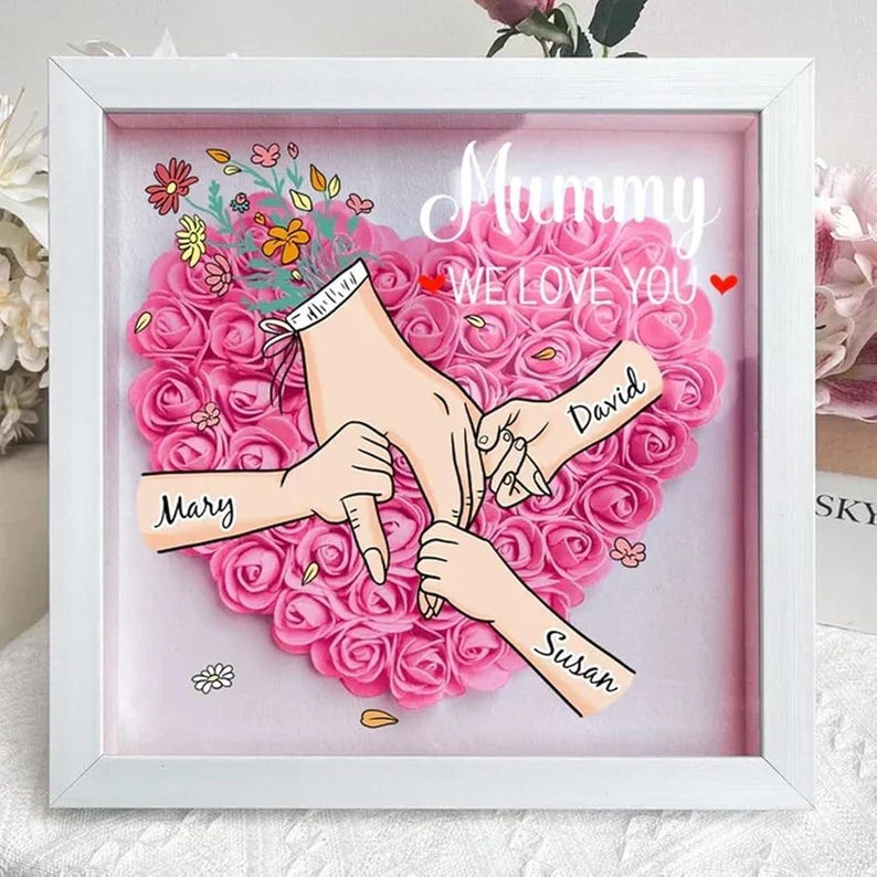 May include: White frame with a pink heart made of roses. The heart has three hands reaching into it, with the words "Mary", "David", and "Susan" written on the arms. The text "Mummy We Love You" is at the top of the frame.